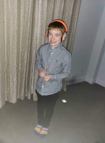 Riley has orange ear defenders, a blue and white checked shirt, black trousers & striped socks. He's smiling & stood in front of a curtain