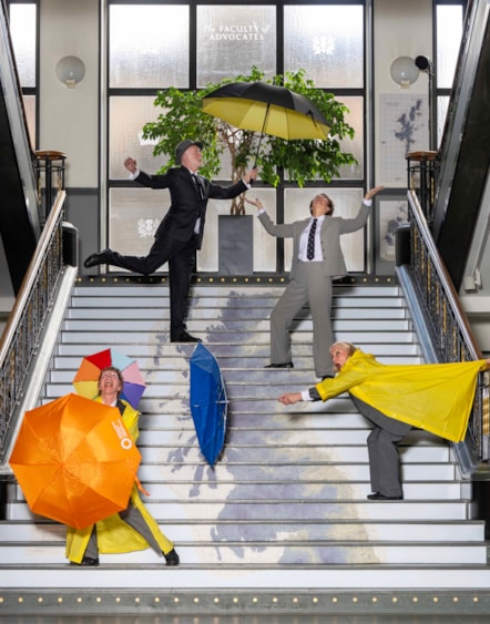A group of dancers dressed in suits and raincoats post with colourful umbrellas on a set of stairs which has a map of Scotland printed on it