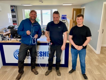 Full length picture of three men standing together holding a sound recording tool that's on a rope and they're smiling towards the camera