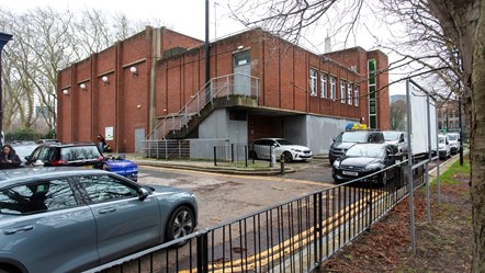 A low rise red brick building with few windows, metal stairs running up one side and metal fire doors, with cars parked outside.