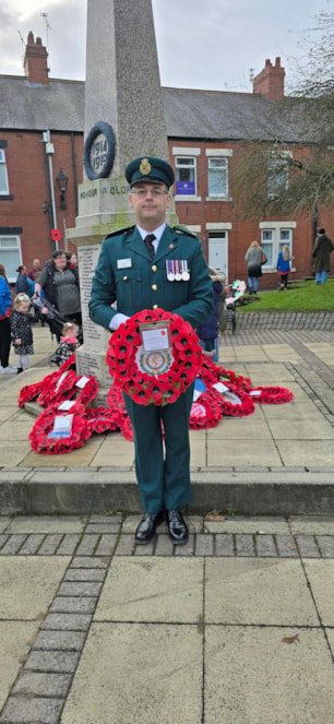 Chris Bradley stood in uniform, holding a poppy wreath with the NEAS badge in the centre,  in front of a remembrance statue.