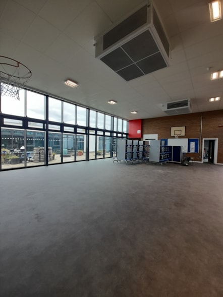 Spacious school hall with grey flooring, large windows, basketball hoops and ceiling-mounted ventilation units.