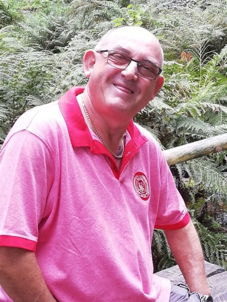 Mike Peacock wearing a pink t-shirt, sitting on a bench with foliage in the background.