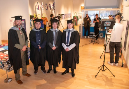 Four males in academic caps and robes stood in front of another group of people recording and filming a podcast