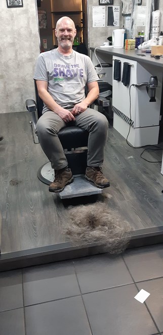 Danny with his head and beard shaven, next to the barbers chair is a pile of his hair