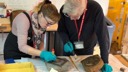 Woman and man examining objects from Lancashire's textile history. They are wearing blue gloves and brushing the objects with paintbrushes