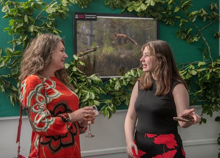 Two females chatting in front of an image of a red squirrel framed upon a green wall dressed with leaves and branches