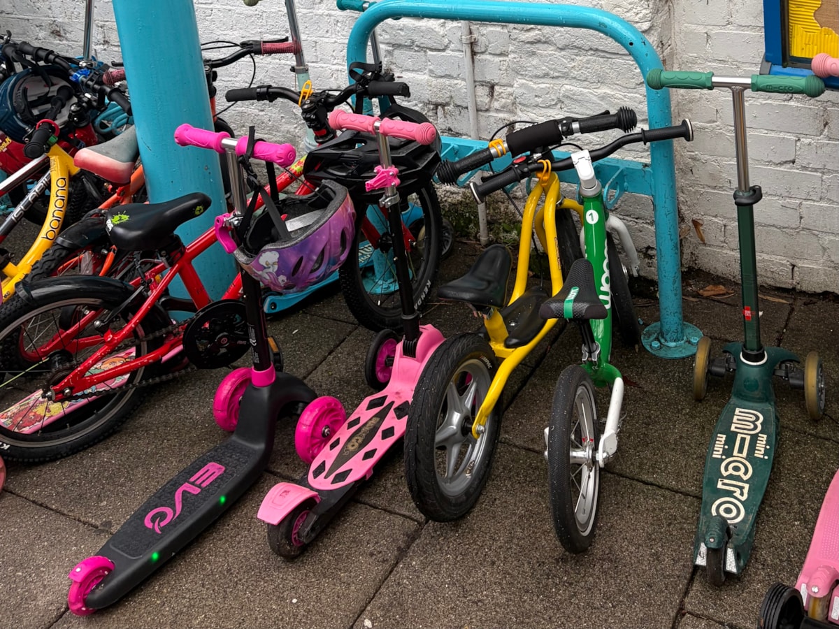A row of colorful children's bicycles and scooters parked against a white brick wall and a blue metal rack