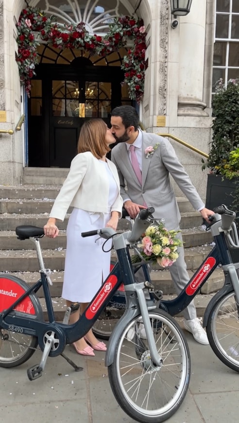 Budding photographers share inspiring images of Santander Cycles to celebrate 15 years of cycle hire in the capital: Nicolas Moura and his wife with Santander Cycles on the steps of Chelsea Old Town Hall surrounded by flowers of their wedding day in 2024