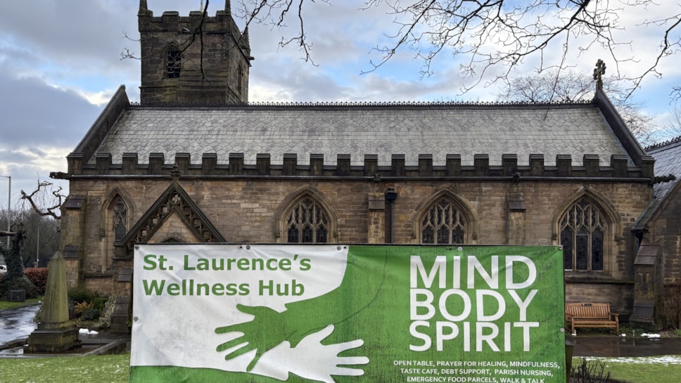 Church building with white and green sign in front that reads St Laurence's Wellness Hub, Mind Body Spirit. Light snow on the ground