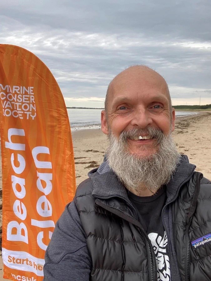 Moray Council's Climate Champion Cllr Draeyke van der Horn at a beach with an orange Marine Conservation Society Flag behind him.