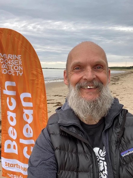 Moray Council's Climate Champion, Cllr Draeyke van der Horn stands at a beach with an orange Marine Conservation Society Flag behind him.