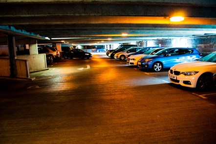 Ground floor of a dimly lit car park with a white arrow painted on the road and seven cars including a white BMW parked on one side.