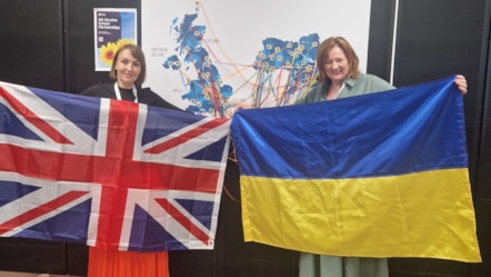 Two women standing side by side indoors, each holding a flag - the Union Jack on the left and the Ukrainian flag on the right.