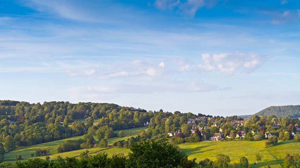 Rural view of gently rolling hills and villages with wooded boundaries in the Cotswolds.