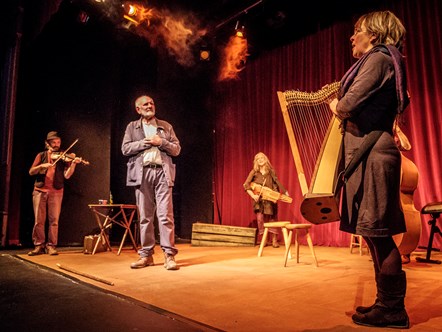 Tom Pow at the centre of the stage with musicians of The Galloway Agreement surrounding playing their instruments with orange lighting