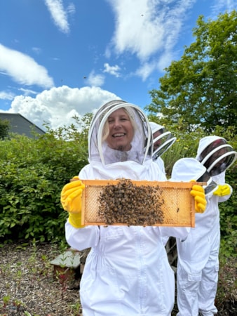 Edinburgh beekeeping 4: Blonde lady smiles at camera and holds up a rack of honey covered in bees. She wears a white bee keeper suit, yellow gloves and mesh face covering. Other staff members stand behind looking at something in the background.