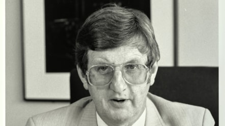 A black and white photo of Colin Ford sat on a chair, wearing glasses and suit.