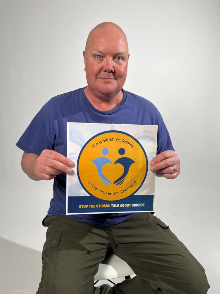 Suicide Prevention Champions - Dominic Brunt: Dominic is pictured sitting on a stool, looking at the camera and holding the Suicide Prevention Champion logo
