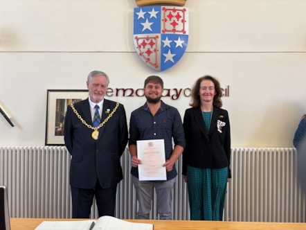 Three people stand in a line smiling at the camera. The person in the middle holds a certificate. There is a coat of arms above them.