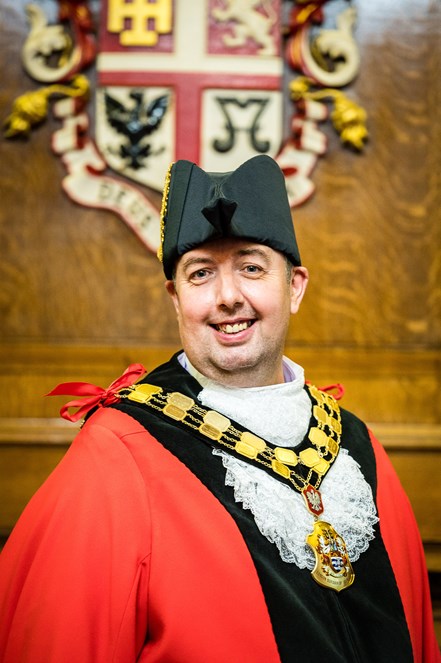 A picture of Cllr Troy Gallagher in Mayoral Robes standing in the Mayor's Parlour in Islington Town Hall in front of the borough crest