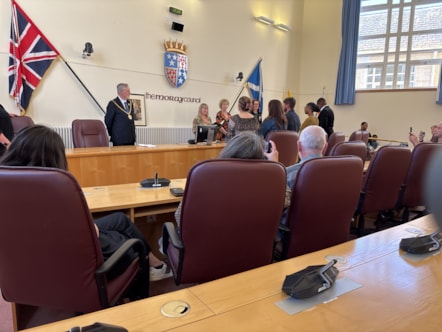 Five people stand in front of a large wooden desk where four officials stand behind. There is a Moray Council sign and coat of arms.