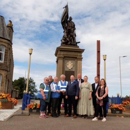 9 people stand against a light blue sky under a war memorial which is crowned by two bronze figures. There are flower planters around.