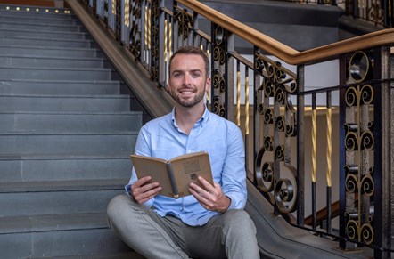 The 2022–23 Scots Scriever, Shane Strachan. He is sitting on stairs and looking at the camera, with an open book in his hands.