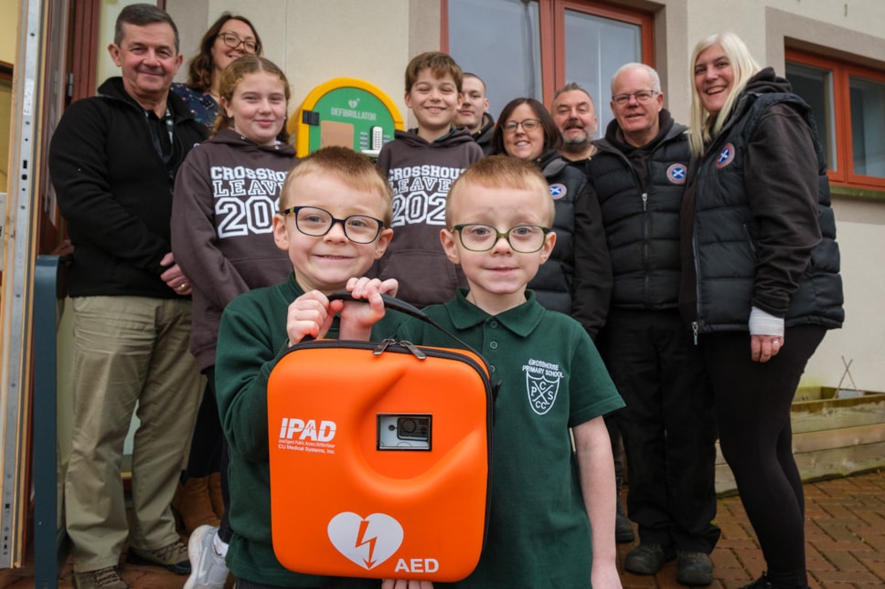 A new defibrillator fitted at Crosshouse Primary School. At the school, Colin McCormack from St John Scotland, Depute Head Kirsty Tomson and the team from Mobityres including Lynne and John Paton. Asha and Reuben from P7 watched over P1 twins Carey and Calvin as they held up the life-saving device