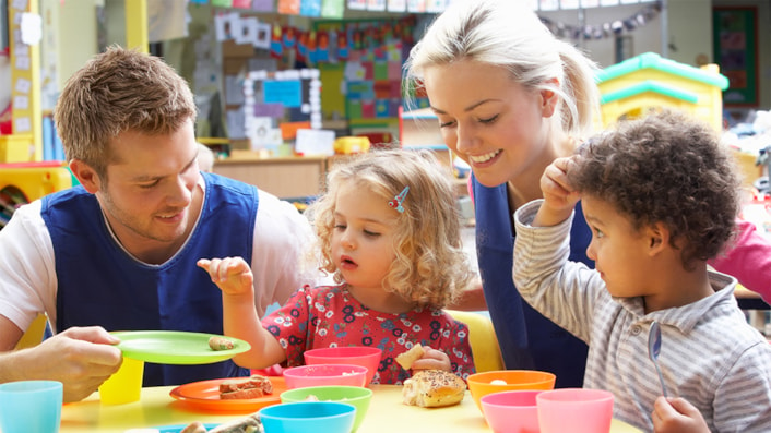 Early years workers sitting at a table with two children and helping them with their lunch.