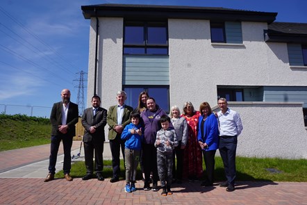 A group of 11 people of mixed ages stand in front of a white two-storey house. The sky is blue and they are standing on terracotta paving.