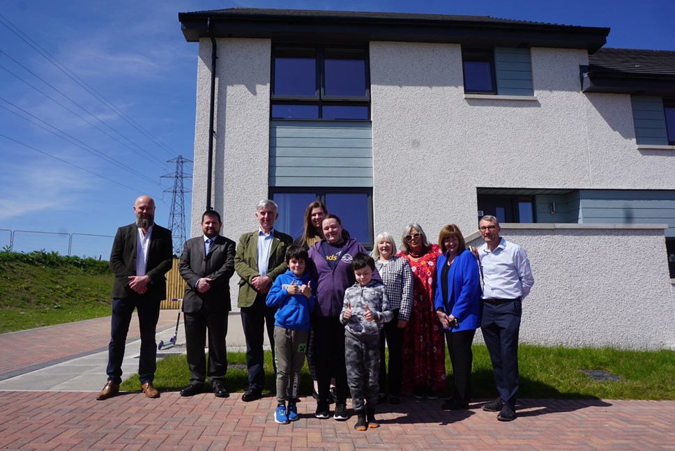 A group of 11 people of mixed ages stand in front of a white two-storey house. The sky is blue and they are standing on terracotta paving.