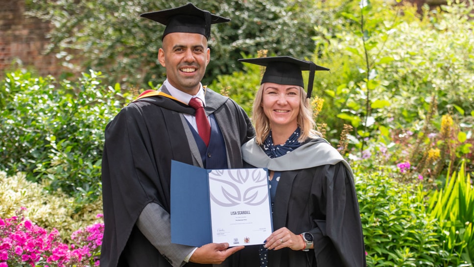 A male and female dressed in academic hats and gowns holding a certificate and stood in front of wildflowers