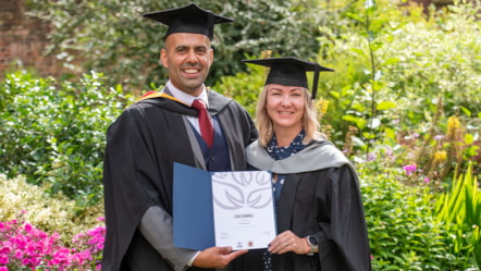 A male and female dressed in academic hats and gowns holding a certificate and stood in front of wildflowers