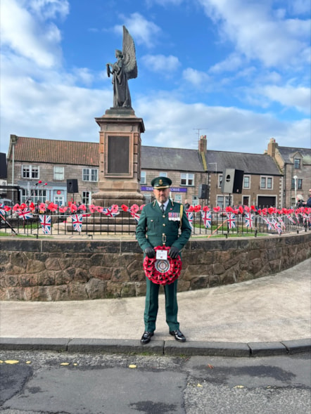 Chris Chalmers in uniform with medals, holding a poppy wreath and standing in front of a statue surrounded by a metal fence bearing poppies.