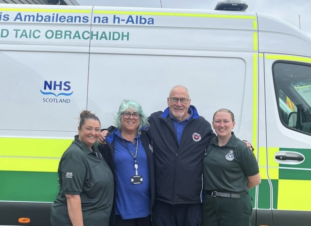 John & Mary: Sam Grieve, Mary and John Kinninmonth, and Laura Cameron standing together, smiling, in front of a parked ambulance vehicle