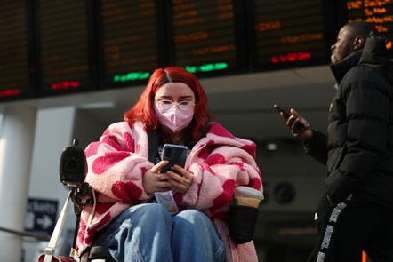A young woman with pink hair sitting in a wheelchair at Birmingham New Street station, looks at the phone she is holding in her hands.