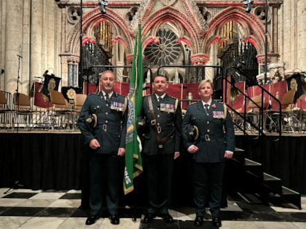 Three people in uniform and medals stood to attention with their caps under their arms in a church holding a NEAS flag pole.