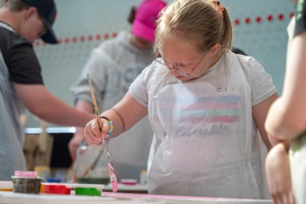 A young girl wearing glasses is painting a piece of wood in front of her