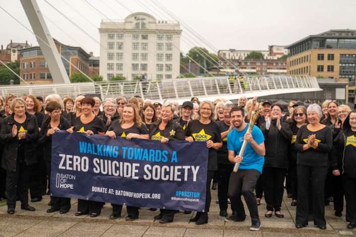 Baton of Hope tour 3: Image shows a crowd of people holding a Baton of Hope banner, alongside a man in a blue branded T-shirt who is holding the baton.