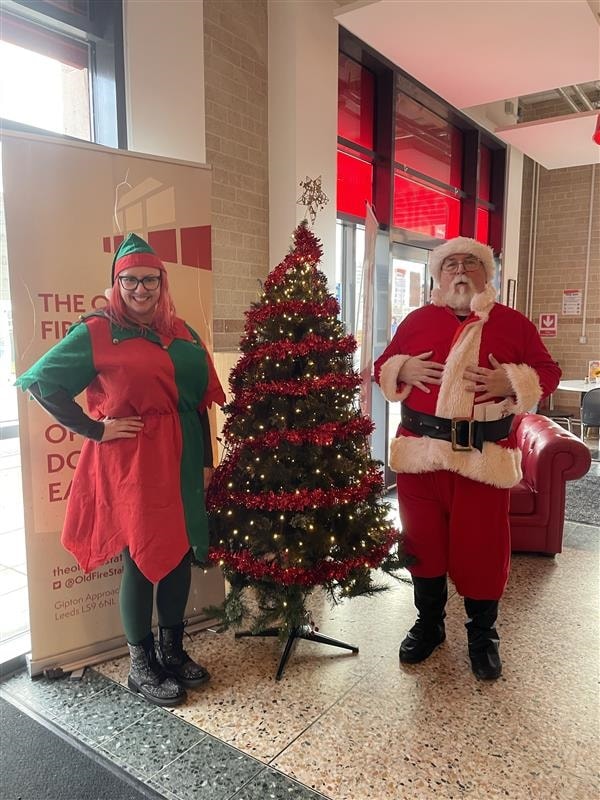 CFL Christmas Gifting 4: Image shows two people dressed as an elf and Santa standing either side of a Christmas tree.