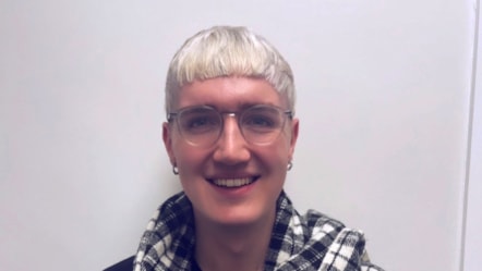Ollie has cropped white blonde hair, wears glasses and a white and black checked scarf on. He's smiling and looking into the camera