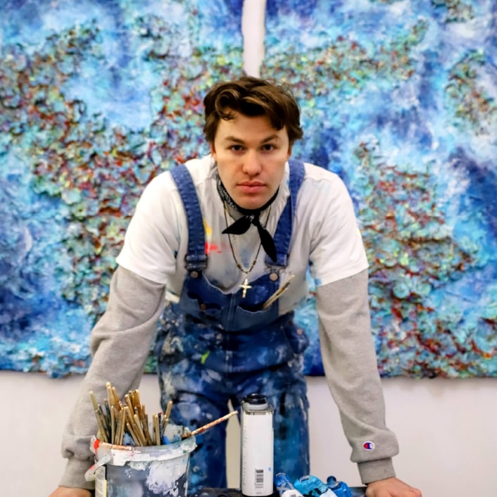Baton of Hope Leeds tour 2: Joshua is looking at the camera, with paint pots and brushes on the table in front of him and blue artwork behind.