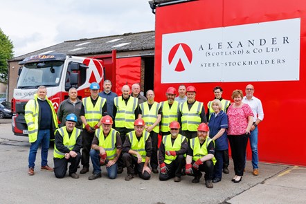 A group of steel workers, some in hard hats, crouch and stand for a group photo in front of a bright red background and company sign