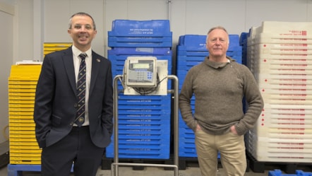 Men are standing next to weighing scales and in front of piles of yellow, blue and white plastic boxes.