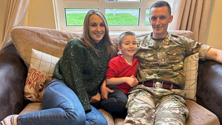 A family of three on a sofa, dad in military uniform, mum in jeans and jumper, young son has red polo shirt on, they are smiling.