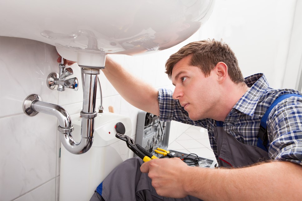 A man in a check shirt with overalls on sits to the right of the picture, facing left underneath a sink. His right hand is on a pipe underneath the sink, he holds a tool in his left hand. An open toolbox sits on the floor in the background.