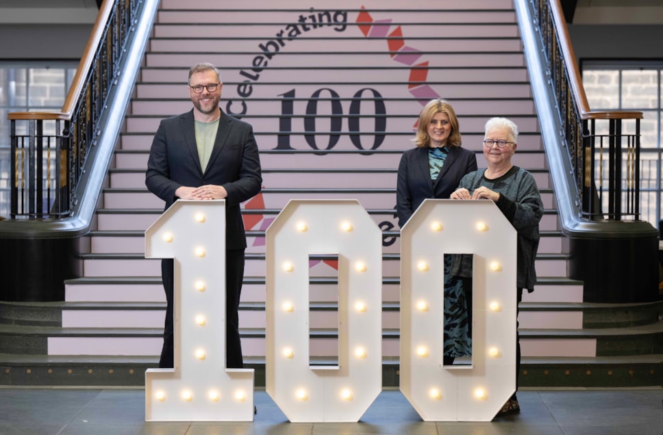 Three people stand in front of stairs. On the stairs, the wording: celebrating 100. The three people stand behind giant numbers '100'.