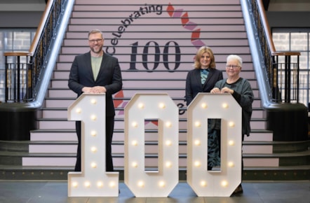 Three people stand in front of stairs. On the stairs, the wording: celebrating 100. The three people stand behind giant numbers '100'.