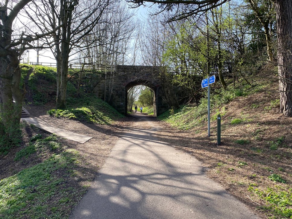 A narrow path leads to a viaduct with two people in the distance on bikes. The sky is blue and there are trees without cover leading the way.